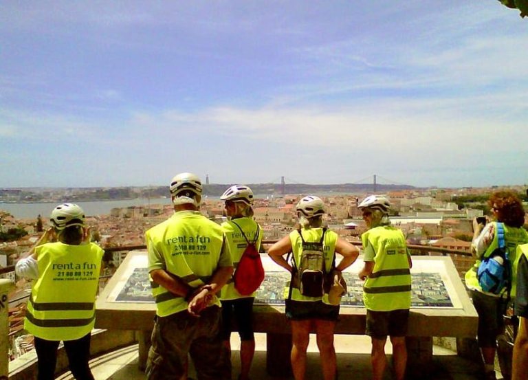 Lisbon electric bike tour. Group in safety vests views city from hilltop. Tagus River and bridge visible. Gay travel advice.