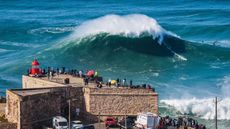 A big wave advances toward the promontory in Nazare, Portugal. There is a small red lighthouse and people watching the wave on the promontory.