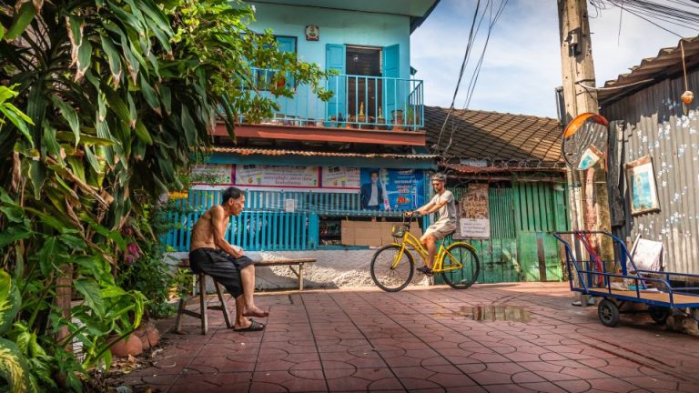 Bangkok Bike Tour: Man on yellow bicycle rides past a local man in a Bangkok neighborhood. Classical bicycle tour for gay men travel advice.