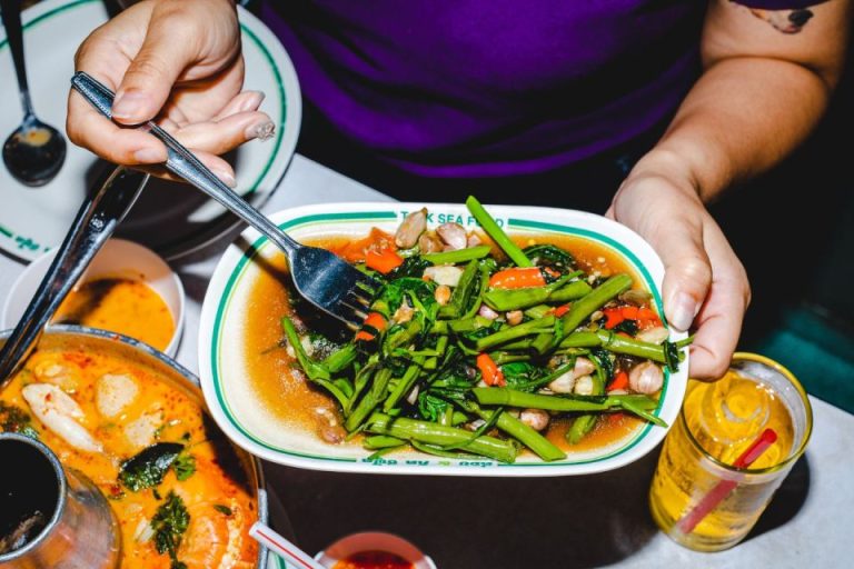 Bangkok Food Tour: Hands hold a plate of stir-fried morning glory, a popular Thai dish. Part of a gay travel food adventure in Thailand.