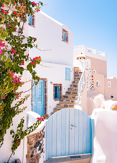 Mykonos gay travel guide: Whitewashed buildings with blue accents, stone steps, and vibrant bougainvillea. A charming Greek island scene.