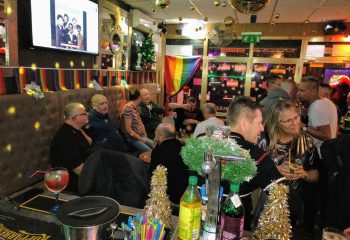 Interior of Betty Boo's gay bar in Benidorm. Men socialize with drinks. Rainbow flag, TV, and festive decor visible. Gay travel advice.