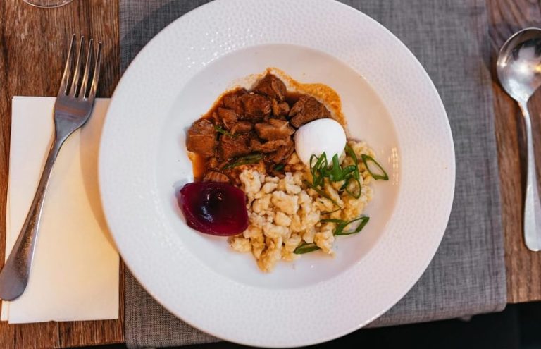 Budapest food tour: A plate of Hungarian cuisine, featuring meat stew, spaetzle, a poached egg, and pickled beetroot. Enjoy gay-friendly travel!