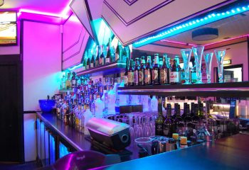 Bar at BJ's White Swan gay club. Shelves stocked with liquor bottles, glasses, and vibrant pink and blue neon lighting.