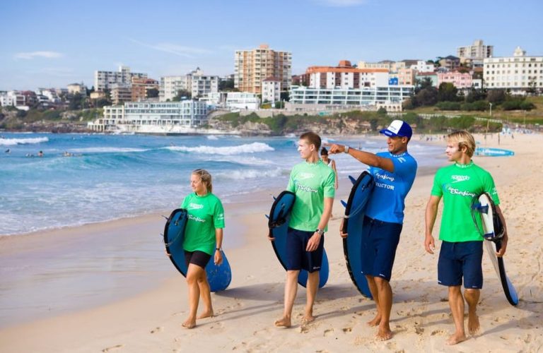Bondi Beach surf lesson. Instructor points to waves with students holding boards. Sydney travel for gay men, beach vacation.