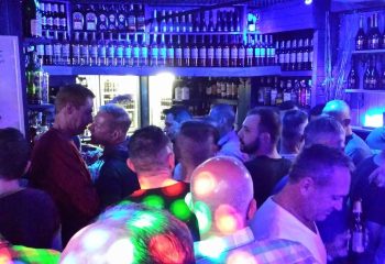 Busy gay bar in Benidorm. Men gather under blue lights, drinks line shelves. Nightlife scene, Brief Encounter gay venue. Travel advice.