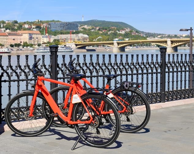 Budapest electric bike tour. Two red bikes parked by the Danube River with Margaret Bridge in the background. Explore the city in style.