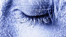 A close-up of a woman's closed eye with blue frost covering her face and eyelashes