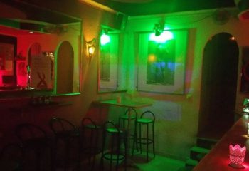 Interior of Chessa Bar, a gay bar in Torremolinos, Spain. Green and red lights illuminate the bar area with stools and artwork on the walls.