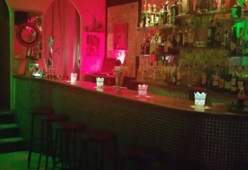 Interior of Chessa Bar, a gay bar in Torremolinos, Spain. Bar stools line the counter with shelves of liquor bottles behind. Red and green lights.
