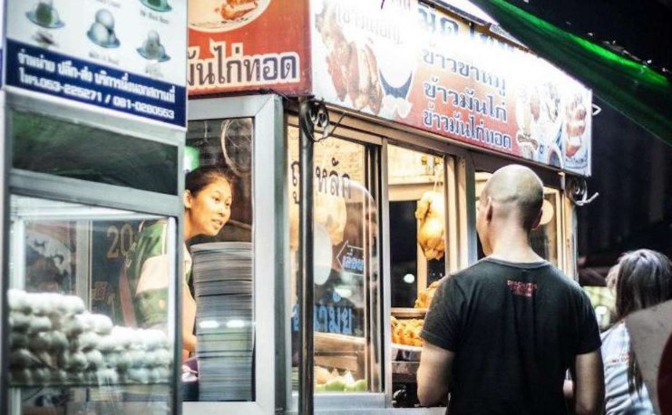 Gay traveler at Chiang Mai street food stall. Woman serves fried chicken. Gay adventure in Thailand. Hanoi Bites tour vibe.