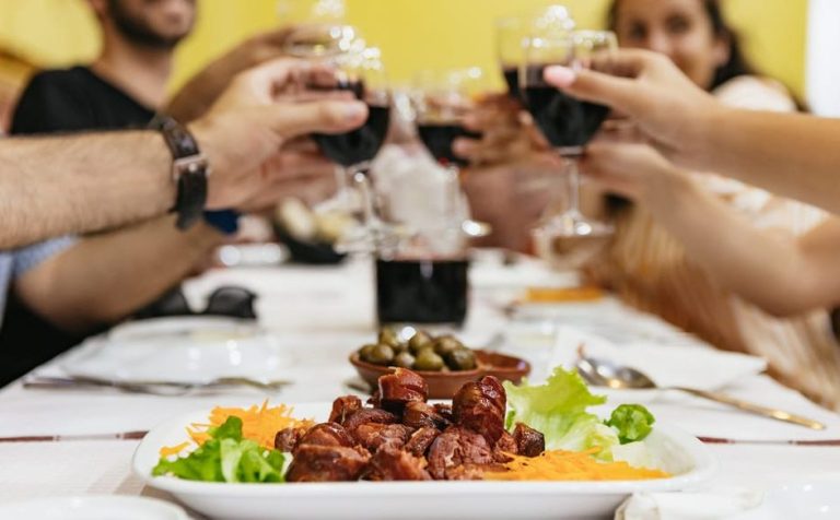 Lisbon food & wine tour: People toasting with red wine during a tasting in Baixa. Chorizo sausage, olives, and lettuce on a white tablecloth.