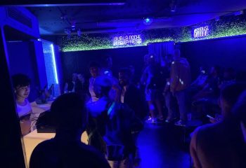 Inside Club XXL, a gay club in Sitges. People dancing under blue lights, with neon signs. Gay nightlife scene, party atmosphere.