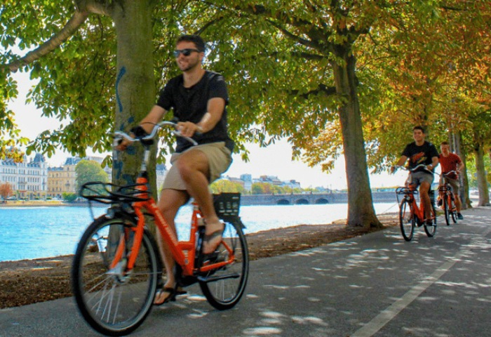 Gay men on a Copenhagen bike tour. 3-hour guided cycle of city highlights. Biking near water, trees, and buildings. Orange bikes.