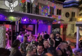 Eagle Bar, a popular gay bar in Torremolinos, Spain. Crowd of men socializing outside the bar, rainbow flags visible.