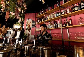 El Gato Lounge gay bar in Torremolinos: Bartender in floral shirt stands behind a bar with bottles, glasses, and floral decorations. Gay travel.