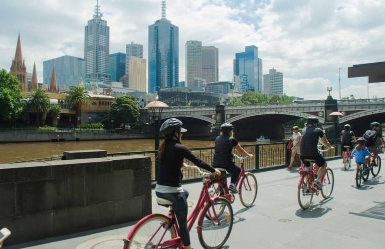 Melbourne electric bike tour. Group cycles along the Yarra River, city skyline backdrop. Explore Melbourne's best spots in style. Gay travel.