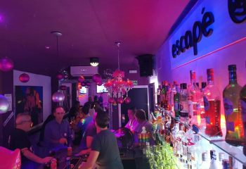 Inside Escape Bar, a gay nightclub in Benidorm. Patrons at the bar, bottles, and purple lighting create a lively atmosphere.