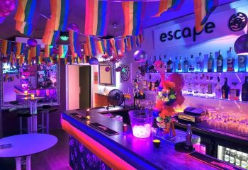 Escape Bar, a gay nightclub in Benidorm. Interior view with rainbow flags, bar with bottles, and neon lighting. Gay travel destination.