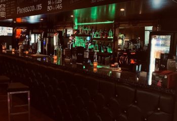 EVA Manchester bar interior. Bottles line the illuminated shelves behind the bar. A sleek, gay-friendly spot for drinks and socializing in Manchester's Village.