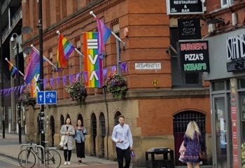 EVA Manchester: Gay travel scene. Rainbow flags adorn a brick building on Richmond St. Pedestrians walk by, showcasing Manchester's vibrant gay culture.
