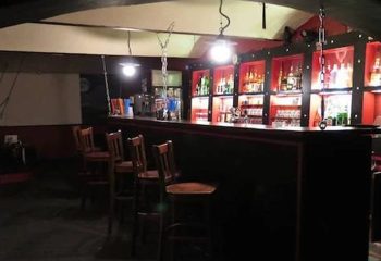 Interior of Factory Club Prague, a gay bar. Dark bar counter with wooden chairs, shelves of liquor bottles, and industrial-style lighting.