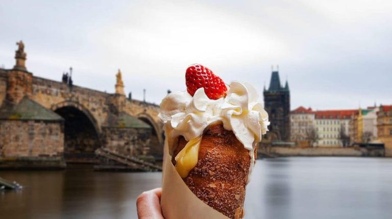 Prague street food: Trdelník pastry with cream & strawberry. Charles Bridge in background. Feast like a local on a guided food crawl.