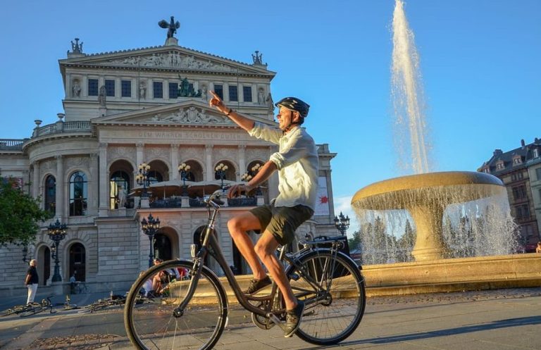 Frankfurt bike tour: Man on bicycle points at Alte Oper opera house near fountain. City's best sights on a gay travel adventure.