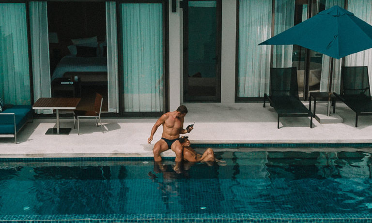 Gay travel: Two men in swimwear relax by a pool at a luxury resort in Thailand. Paradise travel destination for gay men.
