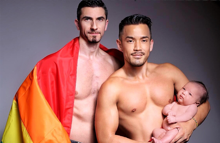 Gay couple surrogacy inspiring story: Two shirtless men, one with a rainbow flag, holding their newborn baby. Family travel advice.