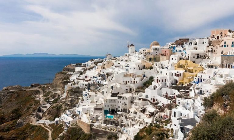 Santorini tour view. Whitewashed buildings cascade down the cliffside. Blue sea and sky. Gay-friendly small group travel.