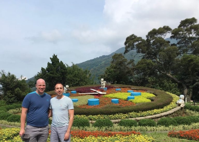 Gay Taipei Private Tour: Two men pose in front of a flower clock in Taipei, Taiwan. Mountains and trees are in the background.