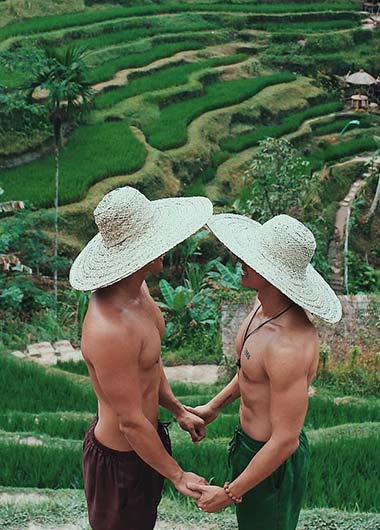 Gay Travel Bali: Two shirtless men in hats hold hands in front of rice terraces. Gay couple enjoying Bali, Indonesia.