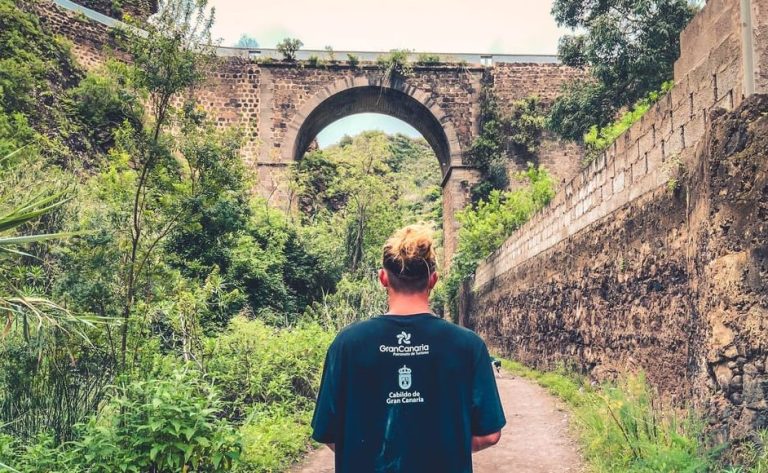Gay man hikes Gran Canaria rainforest tour, a paradise trail. Lush greenery, stone bridge, scenic views. Hike, taste, chill in paradise.
