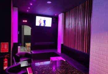 H2O Sauna Benidorm interior. Gay sauna lounge area with purple lighting, seating, TV, and modern decor. Gay travel advice.