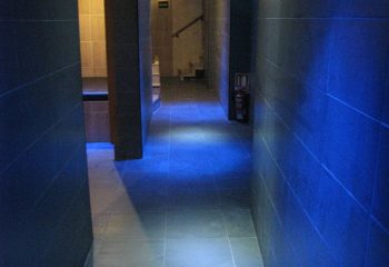 Interior of H2O Sauna, a gay sauna in Benidorm. Blue lighting illuminates the tiled hallway. A fire extinguisher is visible on the right.