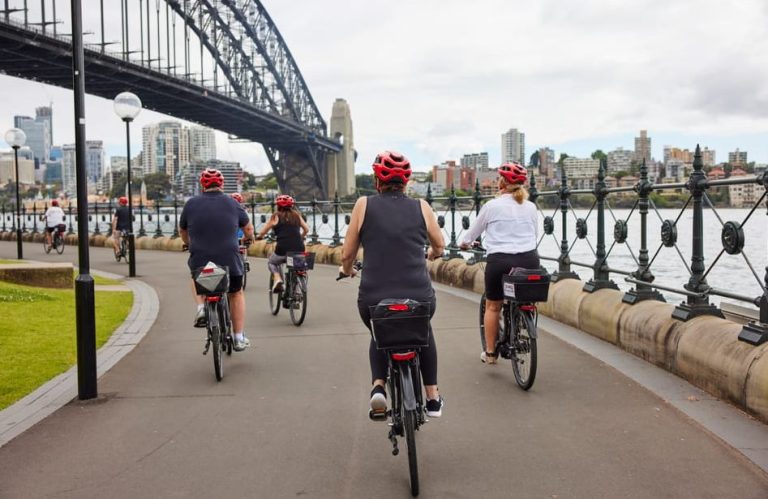 Sydney Harbour e-bike tour. People cycling with Sydney Harbour Bridge and city skyline backdrop. Scenic rides and iconic sights.