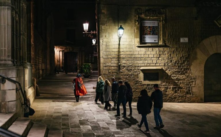 Barcelona's Gothic Quarter night tour. A group follows a guide in a red cape through the haunted streets, exploring legends and ghost stories.