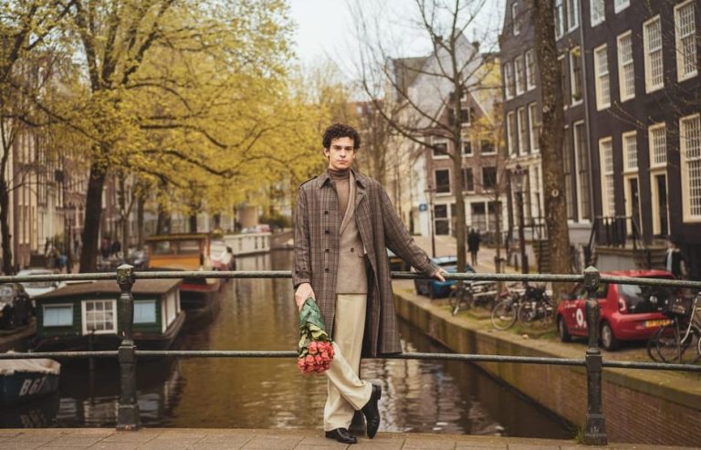 Gay man in Amsterdam photoshoot, canal backdrop. Stylish trench coat, holding roses, perfect Insta shot. Travel advice for gay men.