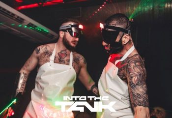 Into the Tank Gay Club Madrid: Two tattooed men in aprons and futuristic eyewear at a party. Nightlife scene.