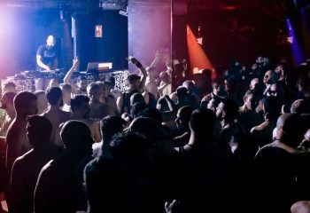 Into The Tank gay club in Madrid: A DJ plays music for a crowd of shirtless men dancing in a dark, crowded space with colorful lights.