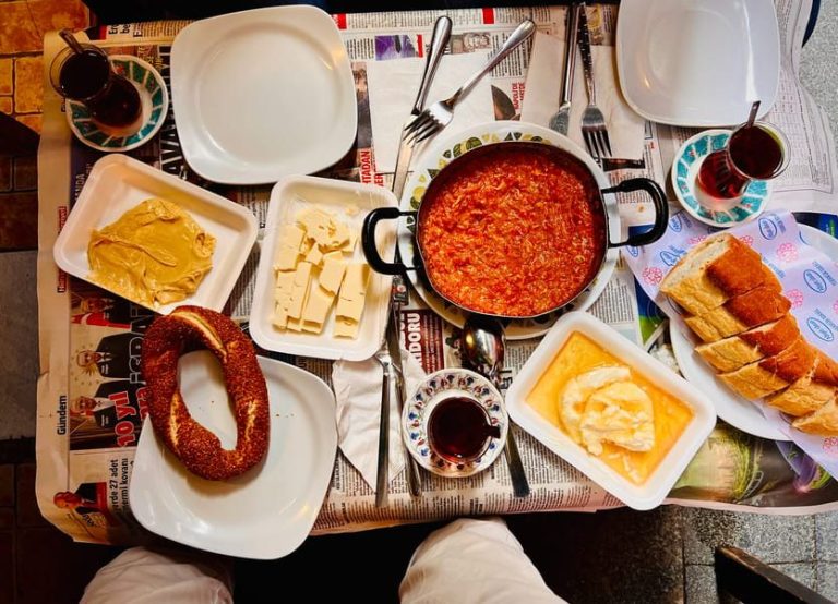 Istanbul food tour: Turkish breakfast spread featuring menemen, simit, cheese, bread, peanut butter, tea. Local eats on newspaper tablecloth.