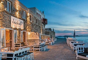 Jackie O' Town Bar in Mykonos. Waterfront view of the gay bar, with outdoor seating and sunset. A popular gay travel destination.
