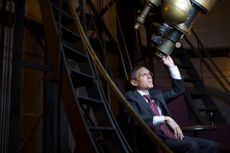 A man in a suit sitting down looks into a large telescope