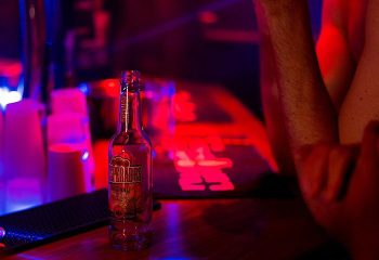 Desperados beer at L'Impact gay bar in Paris. Nightlife scene with red and blue lights, drinks on the bar, and a man's arm visible.