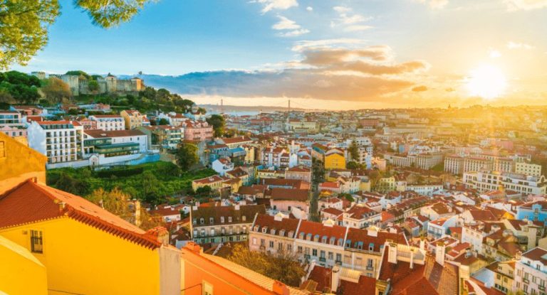 Lisbon's skyline at sunset, featuring colorful buildings and São Jorge Castle. A gay-friendly Tuk Tuk tour explores iconic spots in style.
