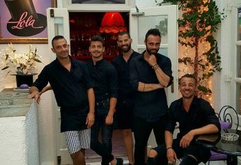 Staff at Lola Bar in Mykonos. Five handsome gay men in black shirts pose at this popular gay travel destination. Nightlife scene.