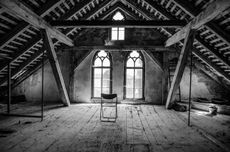 Black and white photo of an empty wooden attic with a metal folding chair in the center.