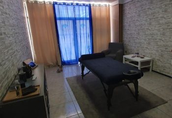 Gay Maspalomas: Massage room in Gran Canaria, featuring a massage table, chair, and a small dog near the window. Cozy spa setting.