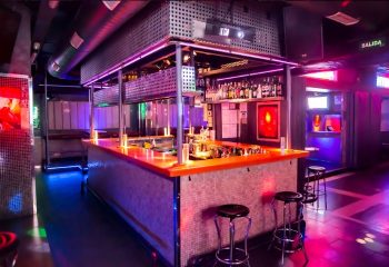 Interior of Mykonos, a gay club in Gran Canaria. The bar is lit with pink and purple lights. Stools are in front of the bar. Nightlife.